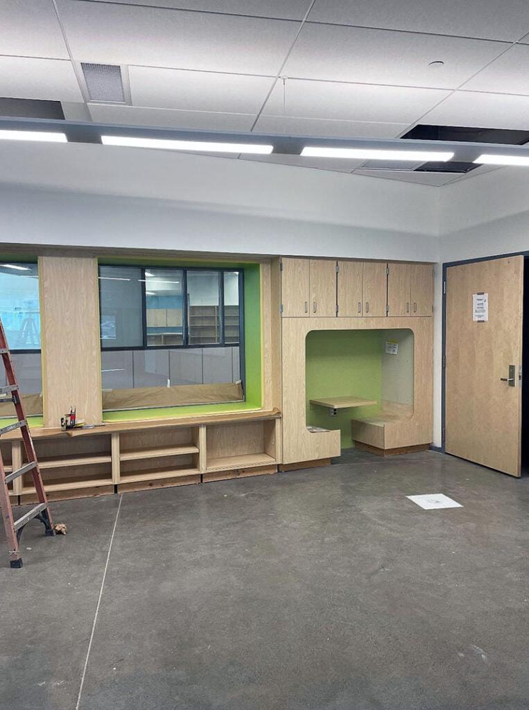 教室的墙壁部分在橱柜下面有一个角落，其中包括两个面向的长凳，它们之间有一个突出的壁架. 旁边是一扇窗户下面的开放式架子. 这两个区域都以明亮的绿色为主调